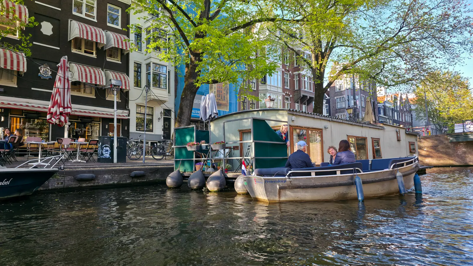 A pale green woonark with flower boxes and a small terrace, as a woman at the window talks to people in a passing boat
