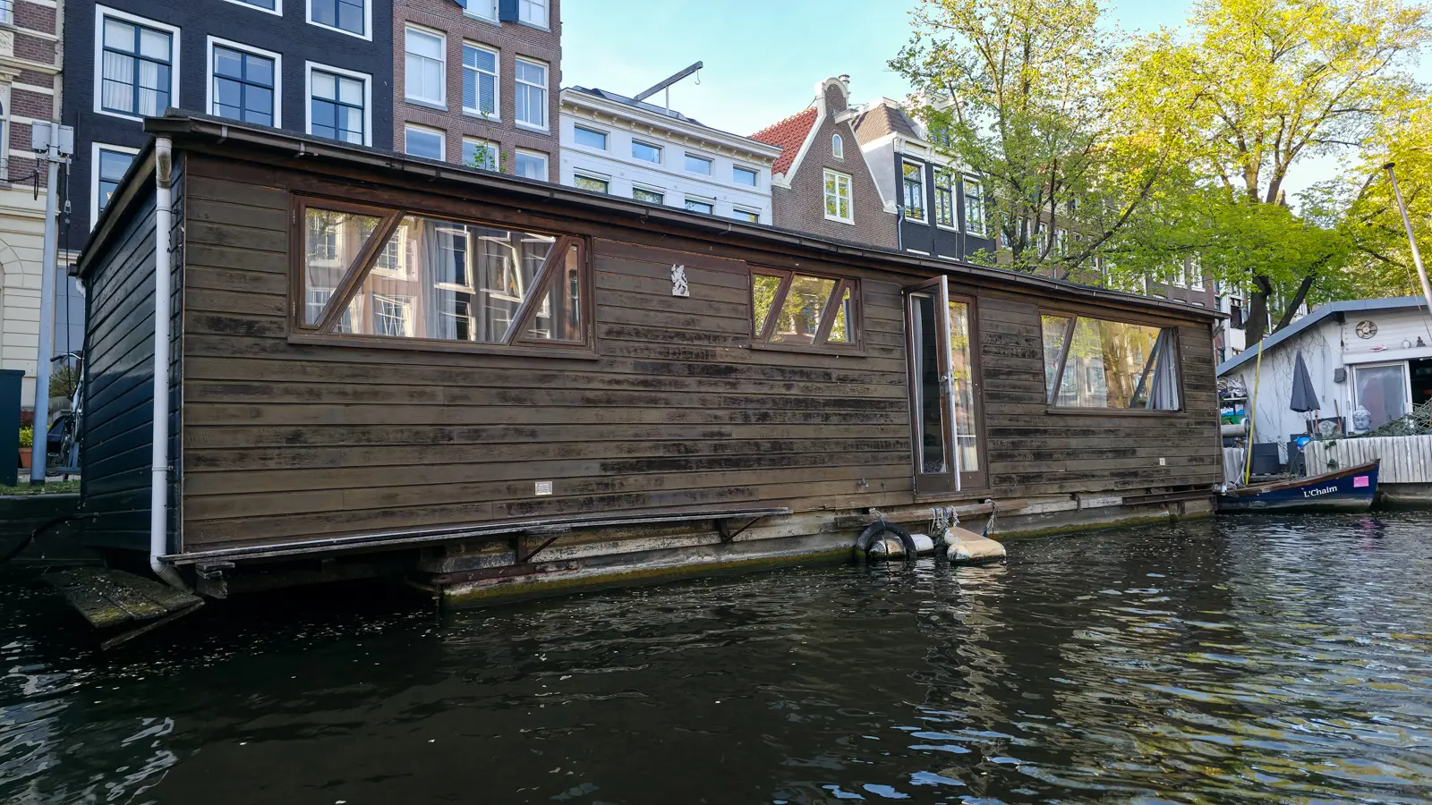A dark wooden woonark with angled window frames and an open glass door, moored in front of gabled canal houses in Amsterdam