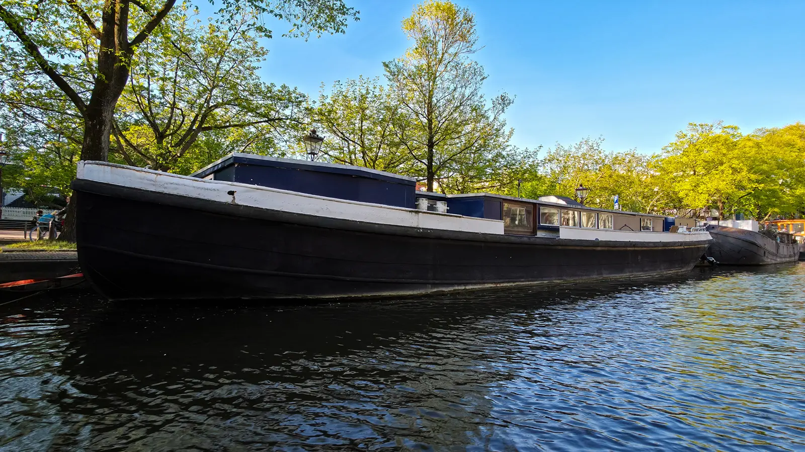 A long black-hulled houseboat with a white gunwale and dark blue cabins, moored under trees on an Amsterdam canal