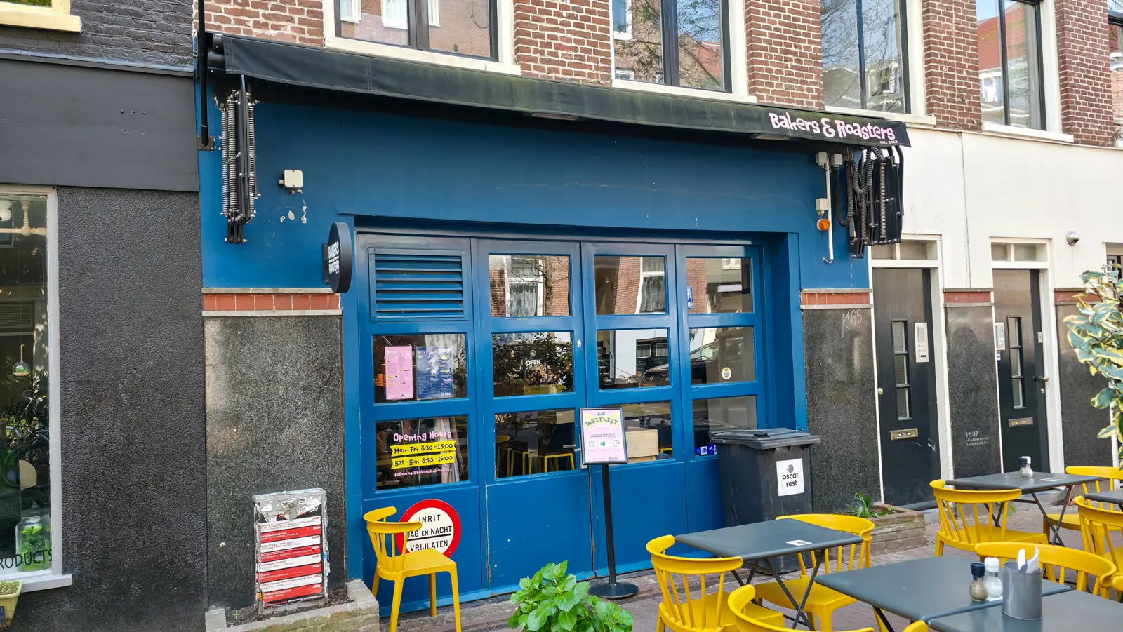 The blue shopfront of Bakers & Roasters with its opening hours painted on the glass and a waitlist sign on a stand outside, De Pijp, Amsterdam