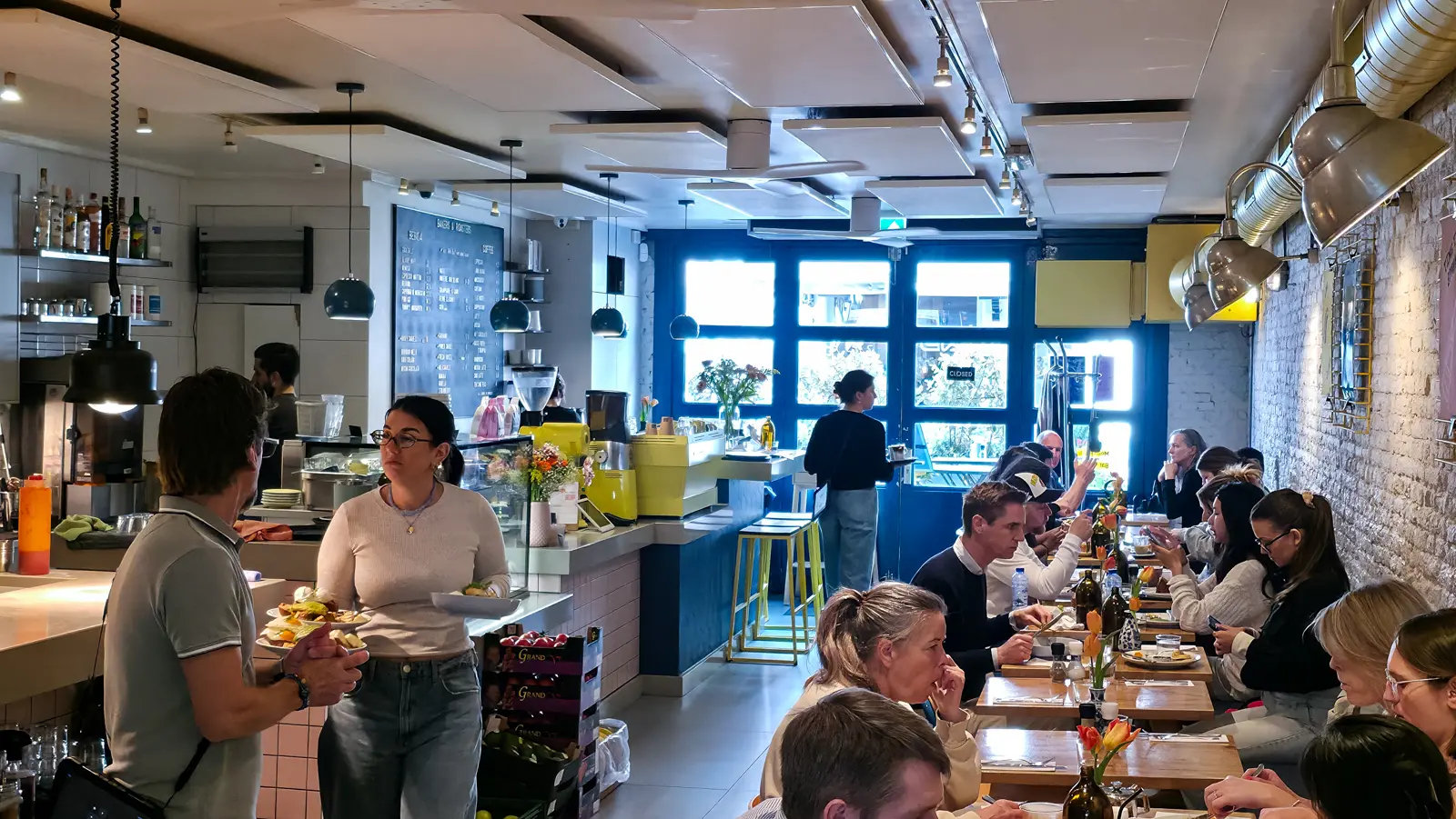 Staff carrying plates past the counter at Bakers & Roasters, with the blue-fronted counter, menu board and full tables against exposed white brick behind