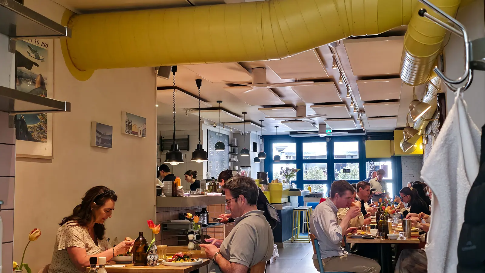 The long narrow dining room at Bakers & Roasters, with a fat yellow ceiling duct running its length, full tables down both sides and a vintage Back to Rio travel poster