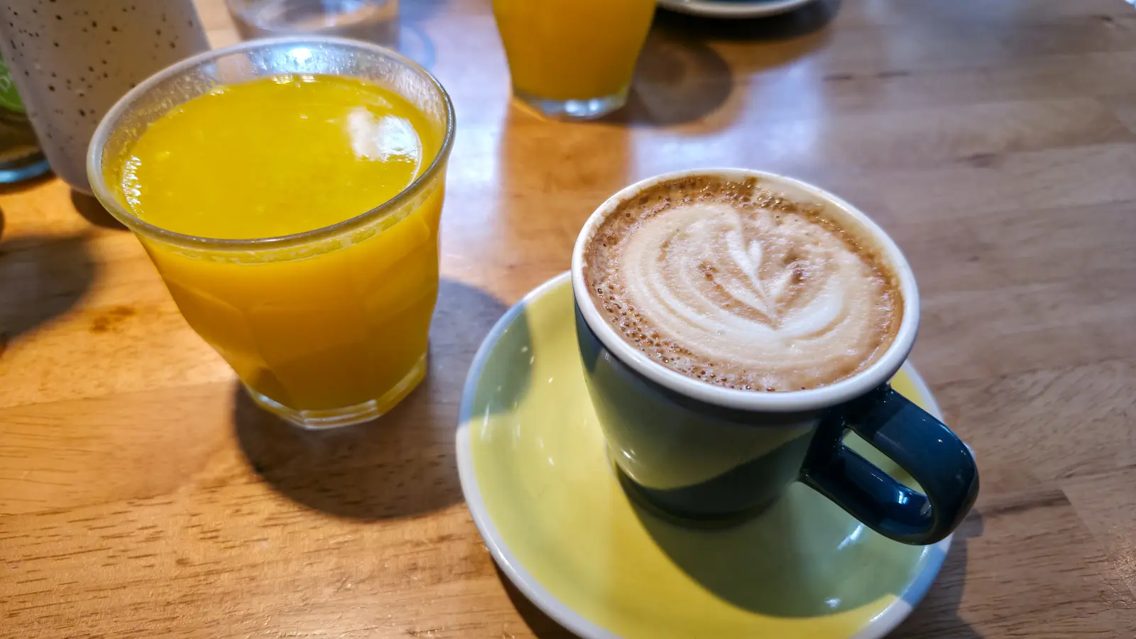 A flat white with a rosetta in a dark green cup on a yellow saucer beside a glass of fresh orange juice on a wooden table at Bakers & Roasters