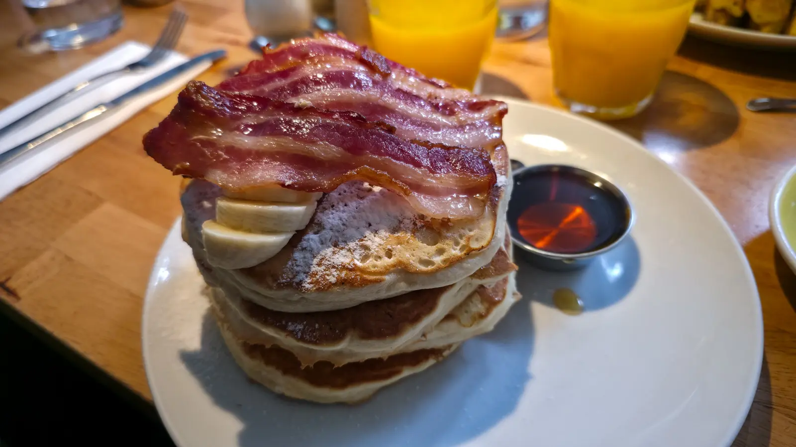 A high stack of five thick pancakes topped with banana, icing sugar and streaky bacon, with a metal pot of syrup, at Bakers & Roasters, Amsterdam