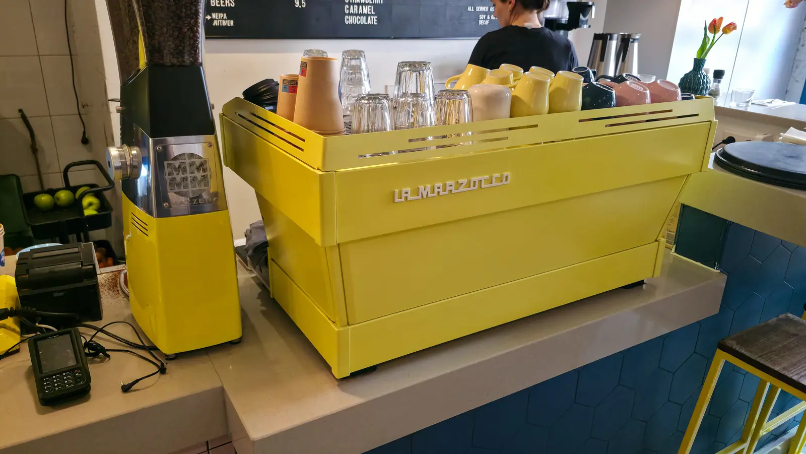 A bright yellow La Marzocco espresso machine and matching yellow Mazzer grinder on the blue-tiled counter at Bakers & Roasters, with cups warming on top