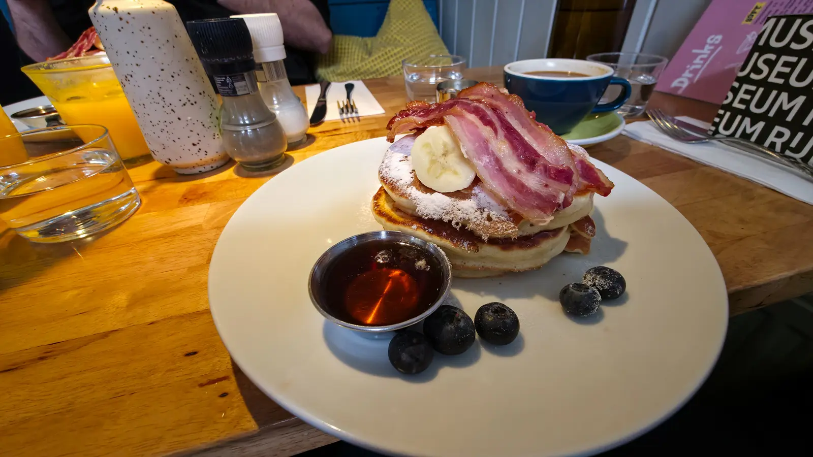 A short stack of three pancakes with banana, bacon and blueberries, a pot of maple syrup and a coffee, on a wooden table at Bakers & Roasters, Amsterdam