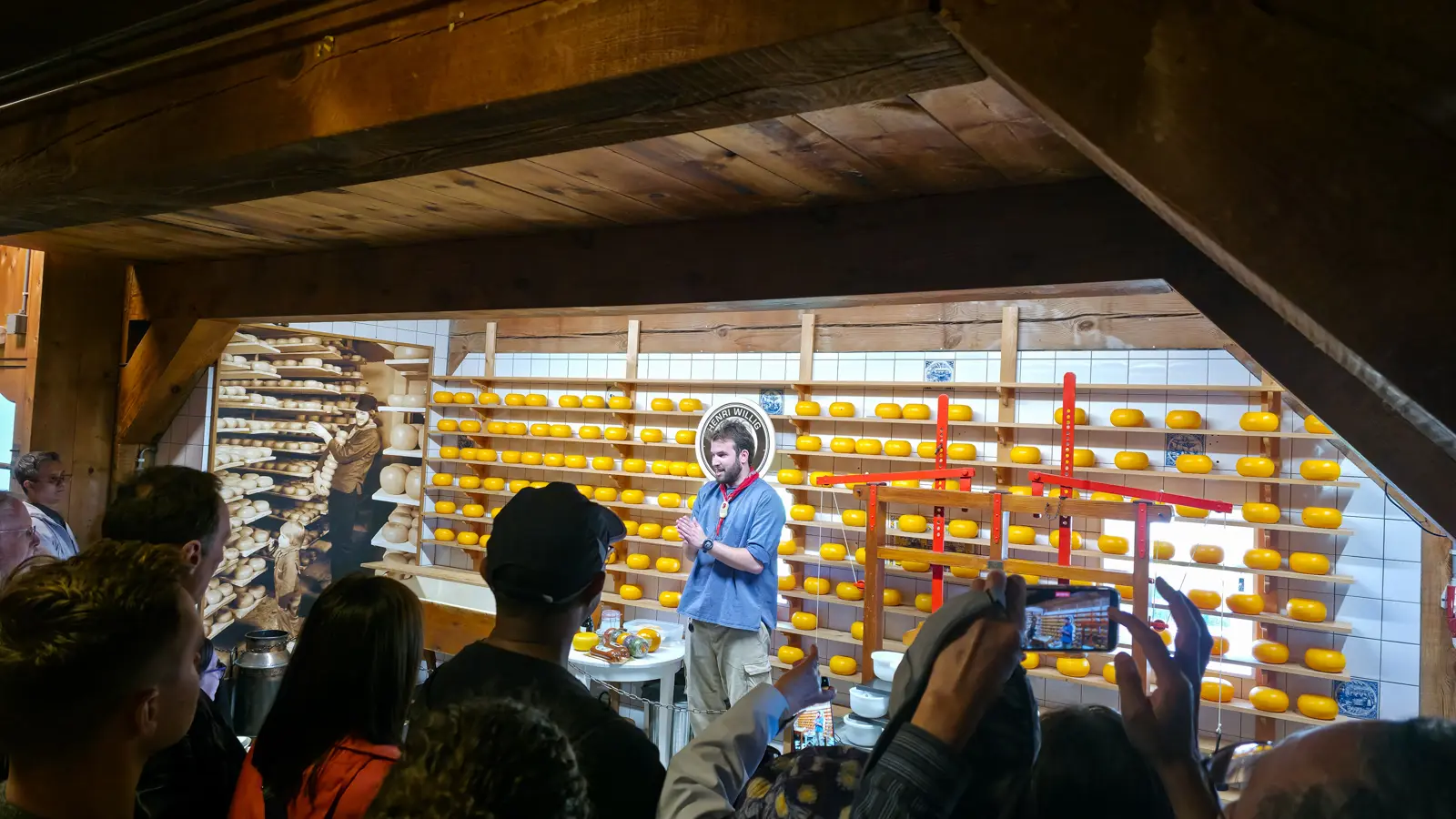 A man presenting the cheese demonstration in front of a wall of yellow wheels and a red press, Catharina Hoeve, Zaanse Schans