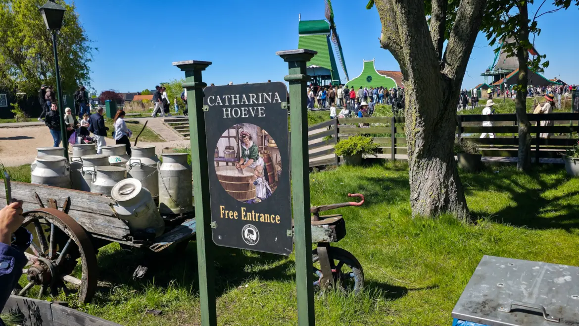 The Catharina Hoeve sign beside a cart of milk churns, with windmills and crowds on the path behind, Zaanse Schans