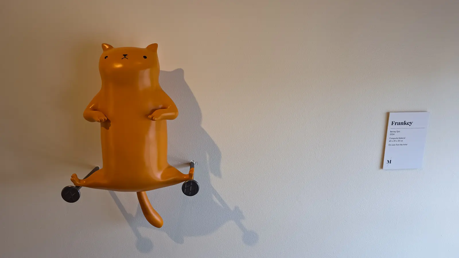 Benney Epic by Frankey, a fat orange cat mounted on its back against the wall with a small black wheel on a rod past each back leg, Moco Museum, Amsterdam