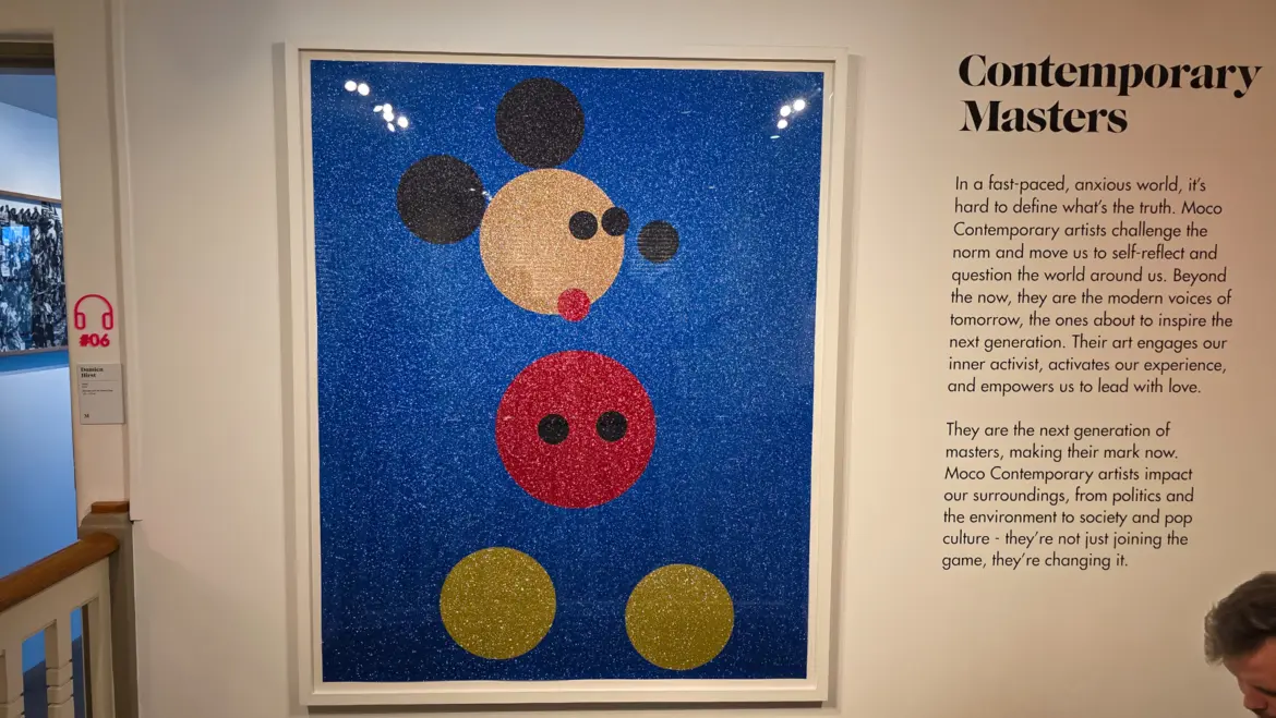 A Damien Hirst glitter Mickey Mouse made of five circles of glitter on a blue ground, beside the Contemporary Masters wall panel at the Moco Museum, Amsterdam