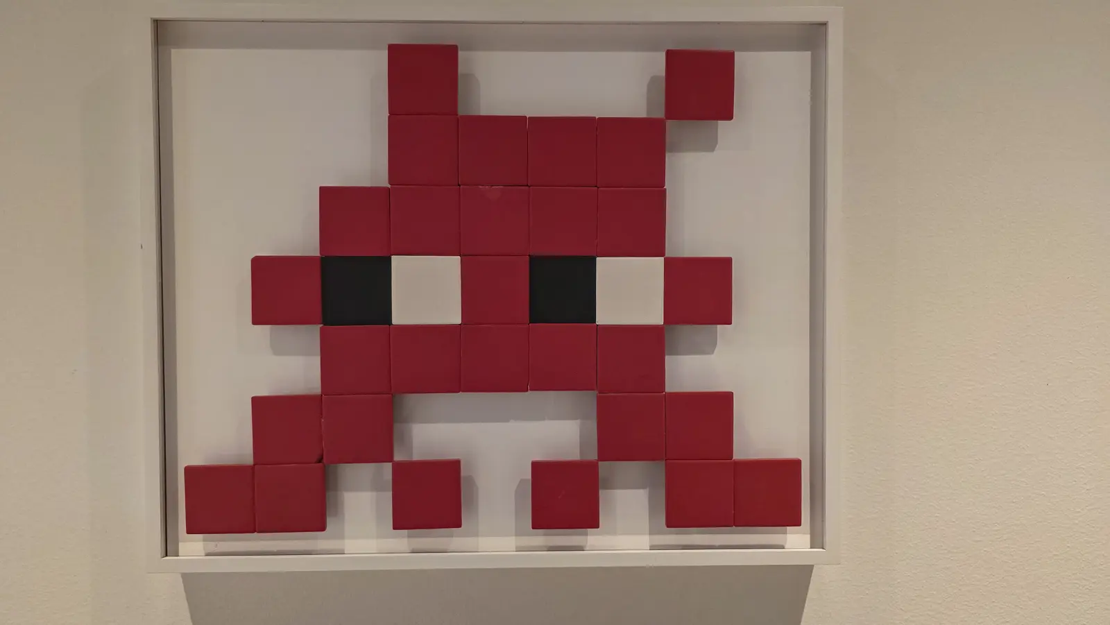 A red and white ceramic tile mosaic by Invader in the shape of a Space Invaders alien, with black and white tiles for eyes, in a shallow white box frame, Moco Museum