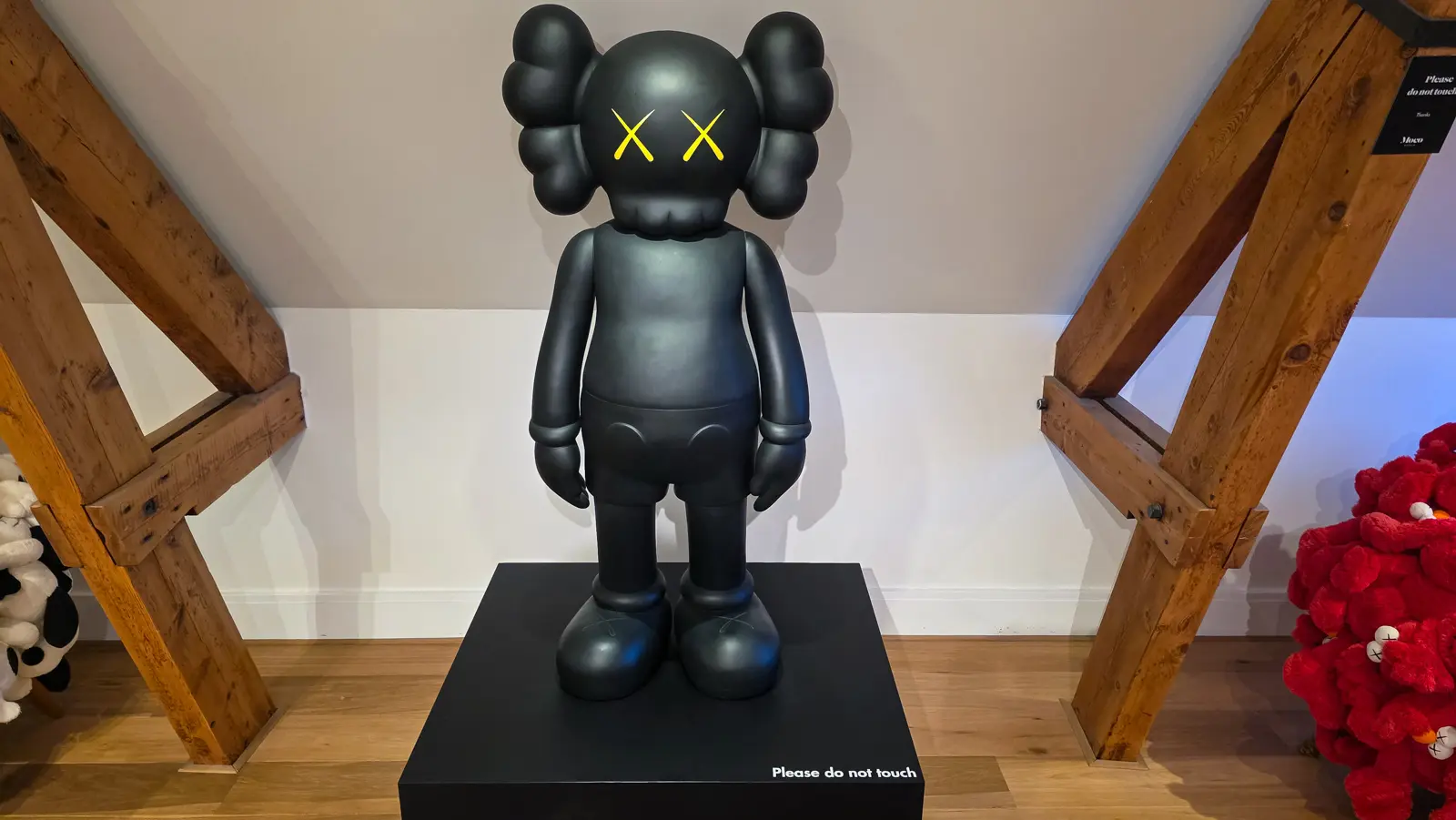 A large matt black KAWS Companion figure with crossed-out yellow eyes and gloved hands standing on a black plinth reading please do not touch, Moco Museum, Amsterdam