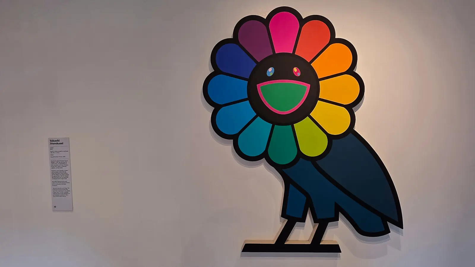 A Takashi Murakami smiling flower with twelve rainbow petals sitting on the dark blue body of an owl, cut out and standing off the wall, Moco Museum, Amsterdam