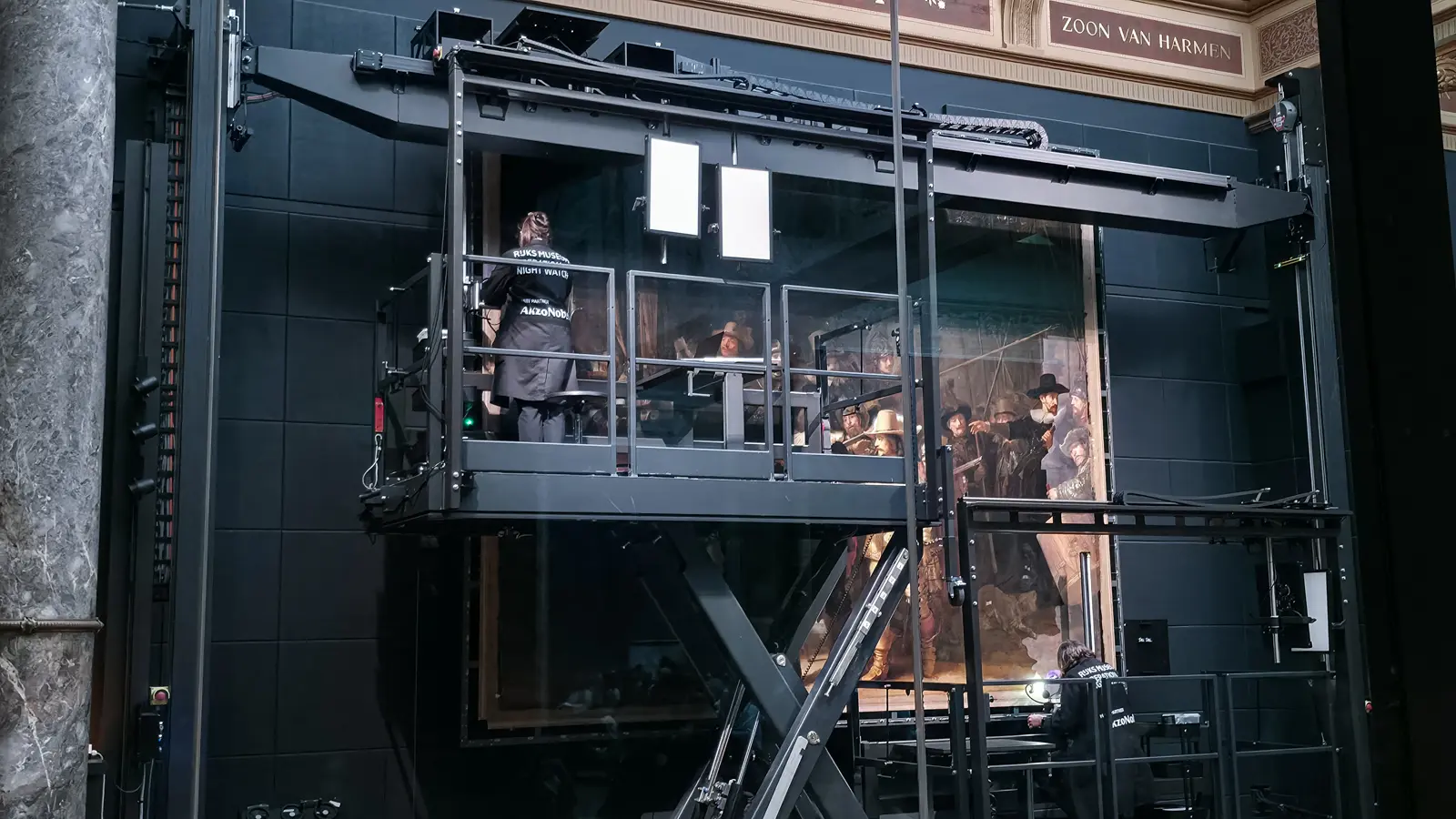 The Night Watch mid-restoration at the Rijksmuseum, Amsterdam, a conservator on the raised platform and gilded lettering above