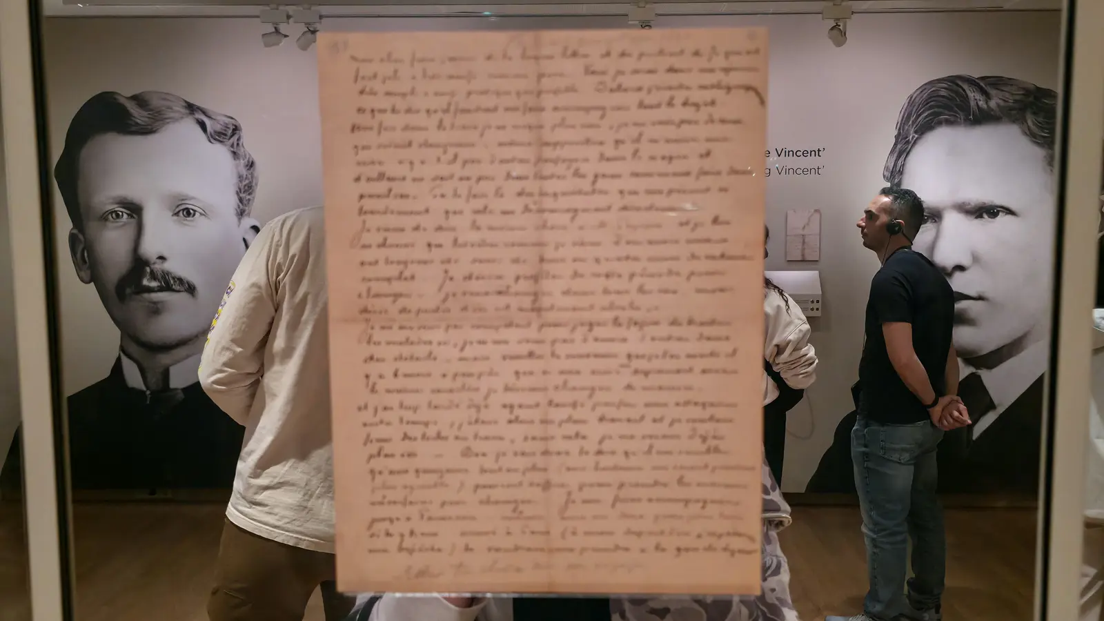 A Van Gogh letter displayed on glass at the Van Gogh Museum, with the wall-height photographs of Theo and Vincent showing through behind it