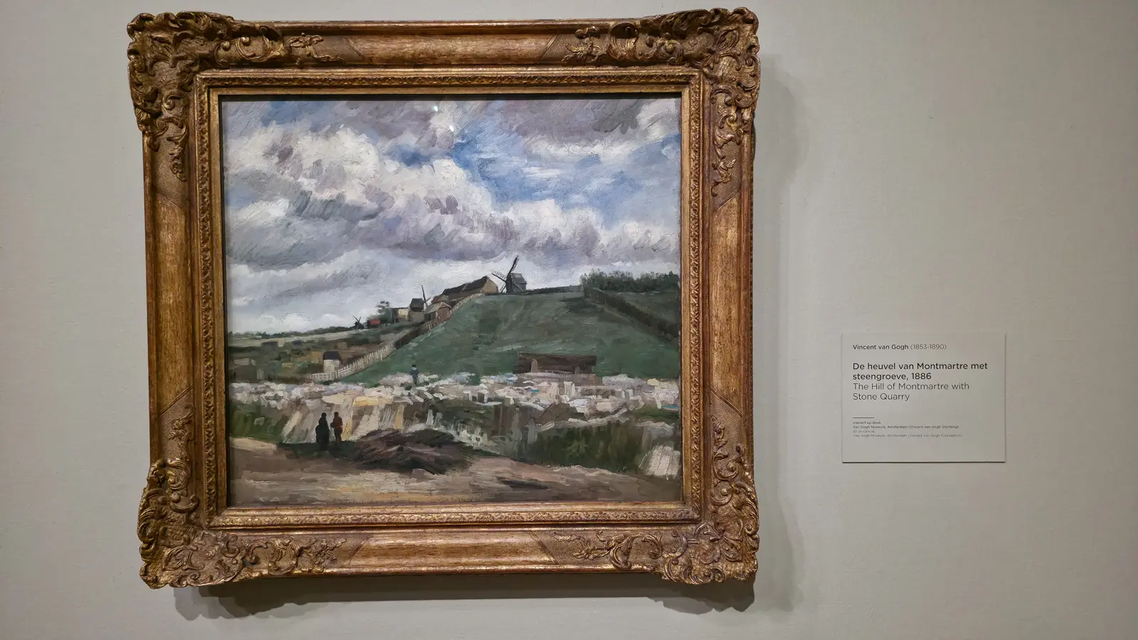 Van Gogh's The Hill of Montmartre with Stone Quarry (1886) in a gilded frame, windmills on the hilltop under a grey sky, with pale quarry blocks and two small figures