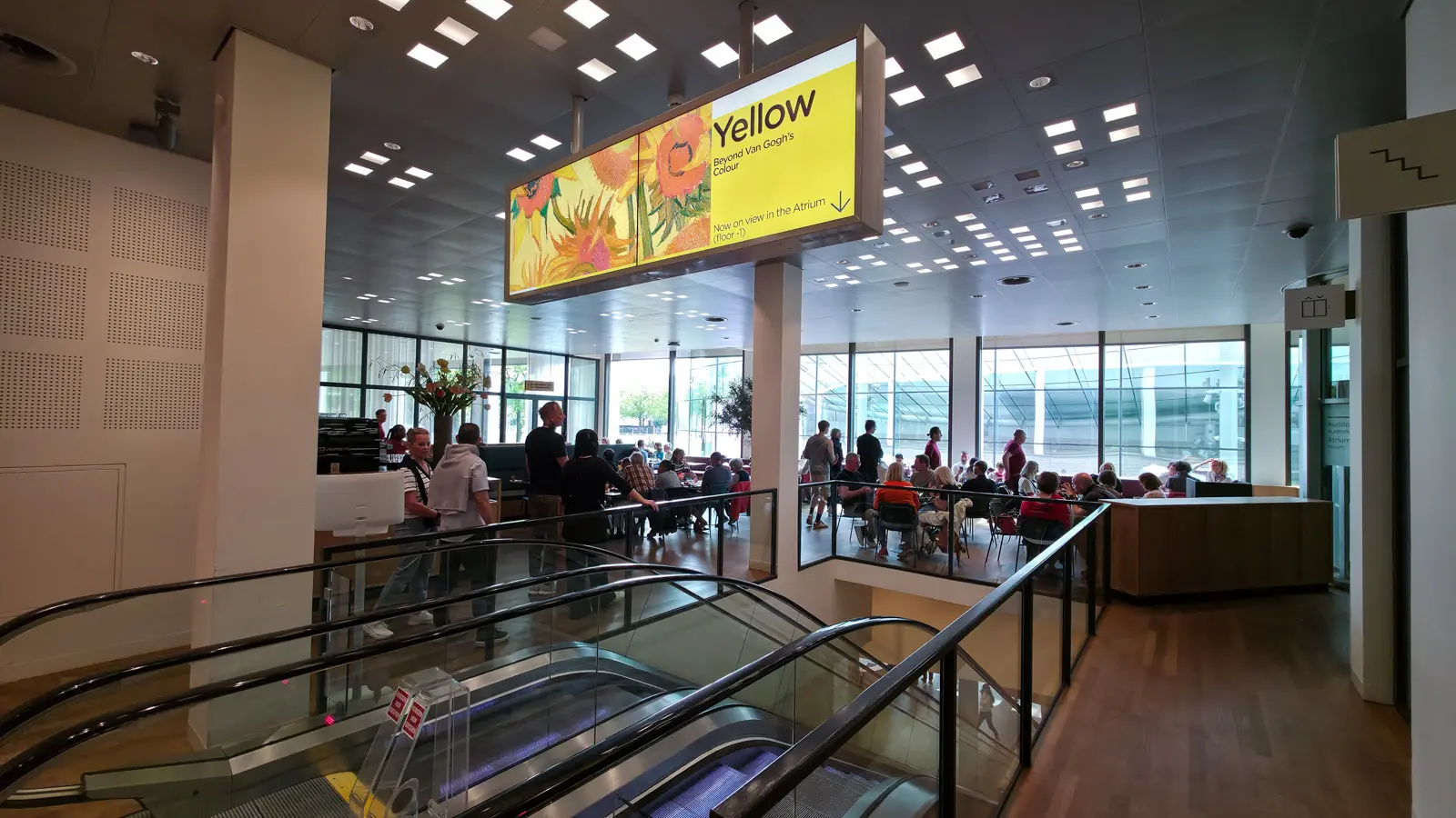 Escalators inside the Van Gogh Museum, Amsterdam, below an illuminated sign for the Yellow exhibition, with café seating visible beyond