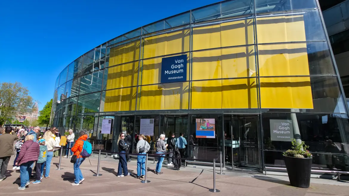 Curved glass facade of the Van Gogh Museum on Museumplein, Amsterdam, a yellow banner behind it and visitors queueing at the doors
