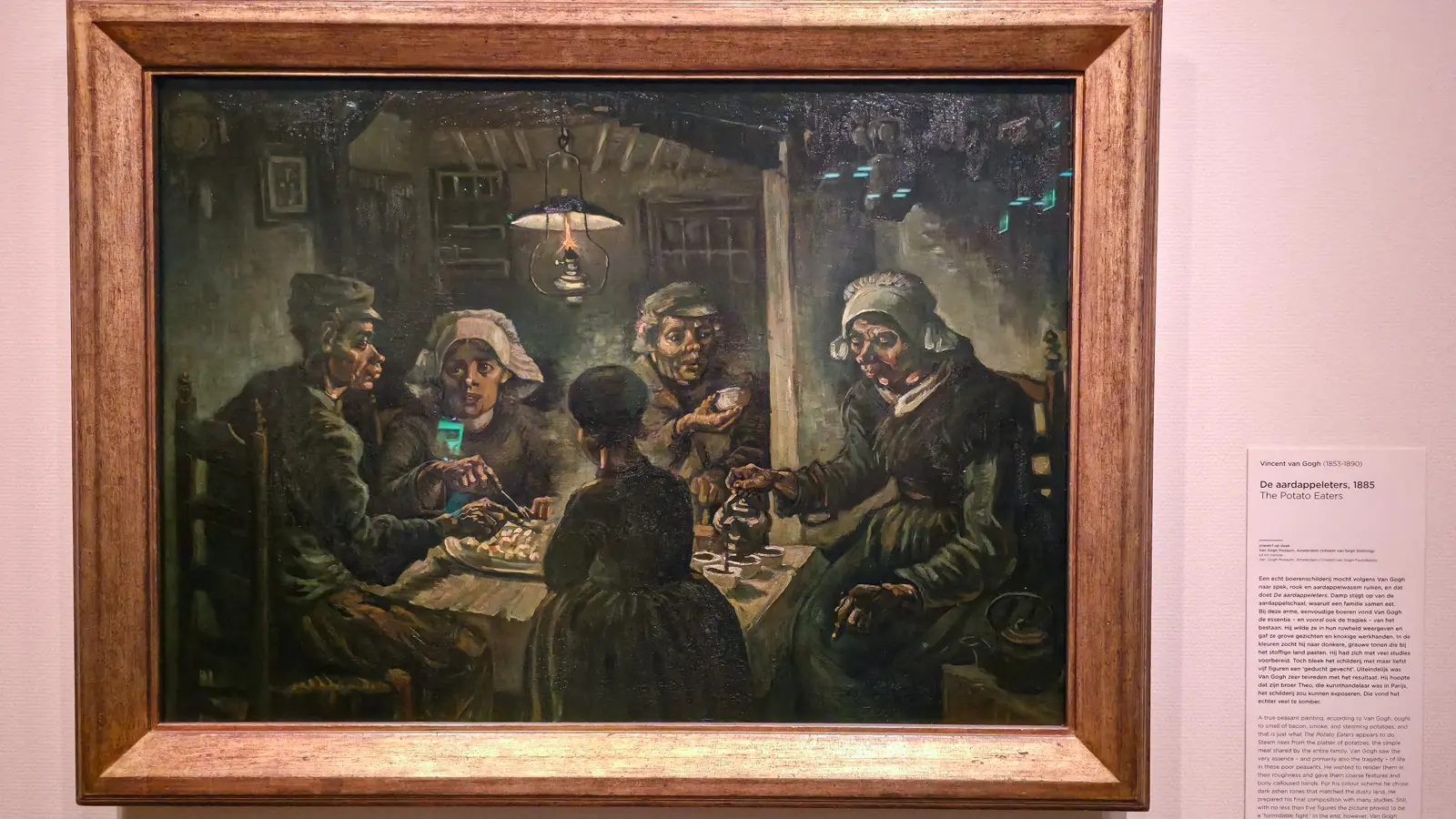 The Potato Eaters by Vincent van Gogh, 1885, in its frame on a pale wall at the Van Gogh Museum, Amsterdam, with its wall label alongside