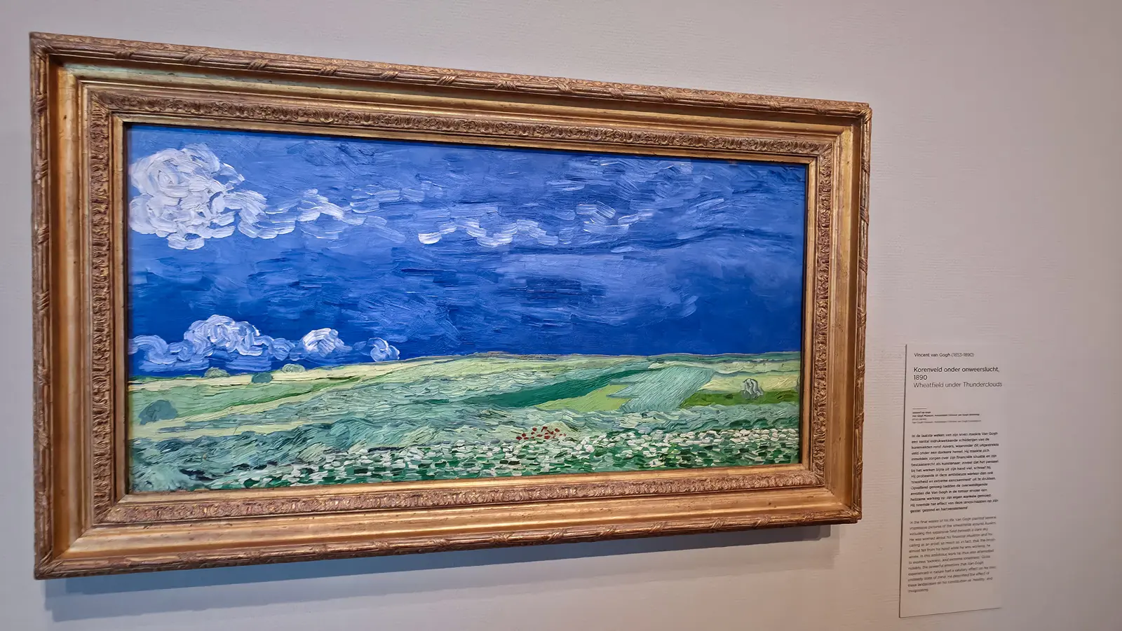 Van Gogh's Wheatfield under Thunderclouds (1890), a wide green field under a deep blue sky with thick white clouds on the left, in a gilt frame, Van Gogh Museum
