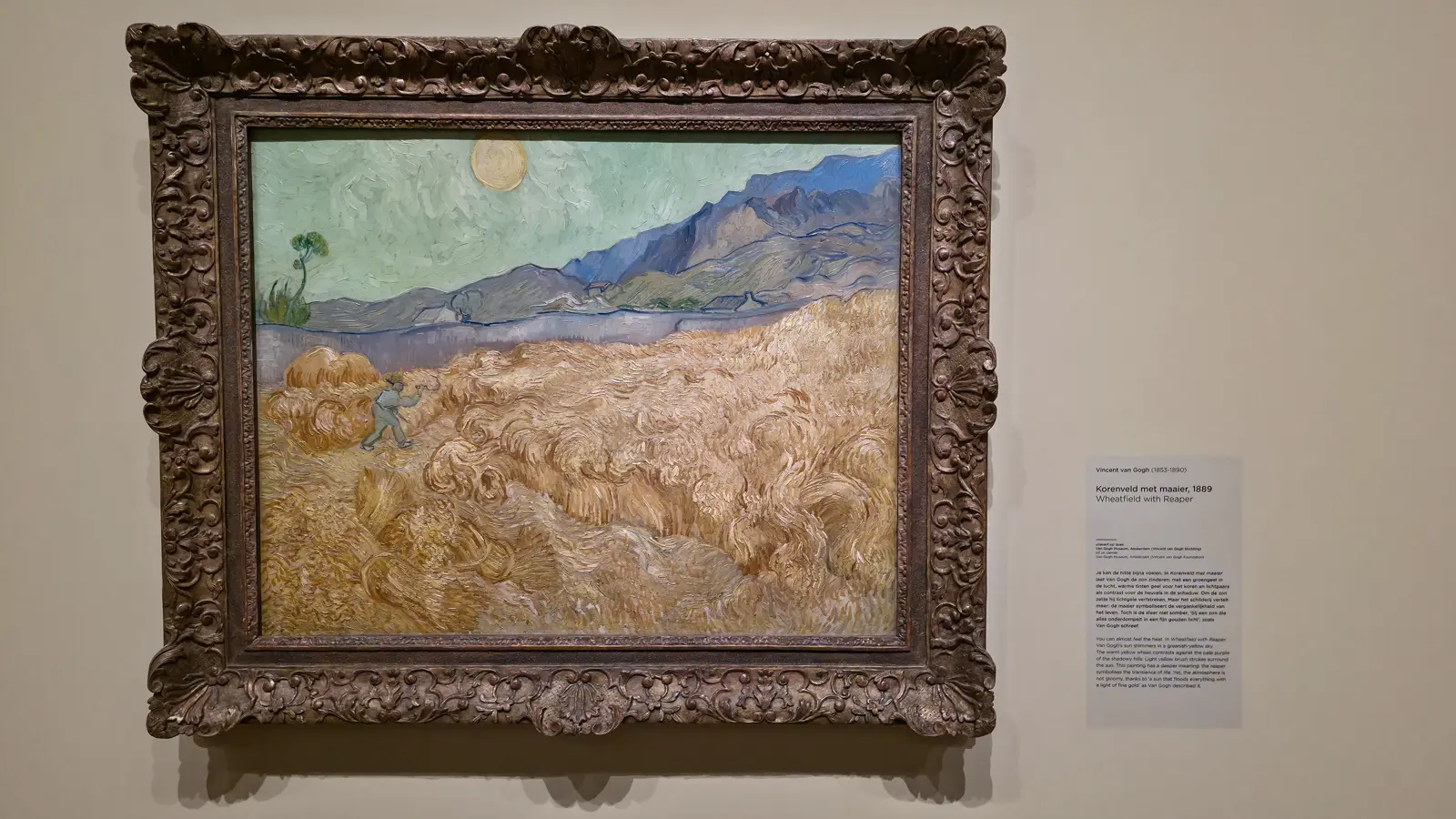 Van Gogh's Wheatfield with Reaper (1889), a lone figure cutting golden wheat under a pale sun and purple hills, in an ornate frame at the Van Gogh Museum