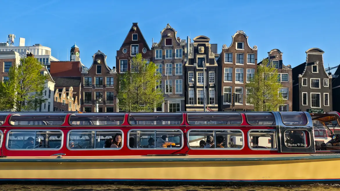 A red and cream glass-roofed canal boat passing a row of gabled houses on the Amstel in Amsterdam under a clear blue sky