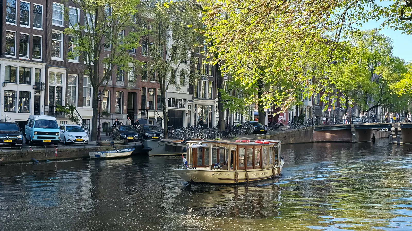 A Flagship Amsterdam saloon boat on a tree-lined canal, with parked bikes, canal houses and a bridge behind