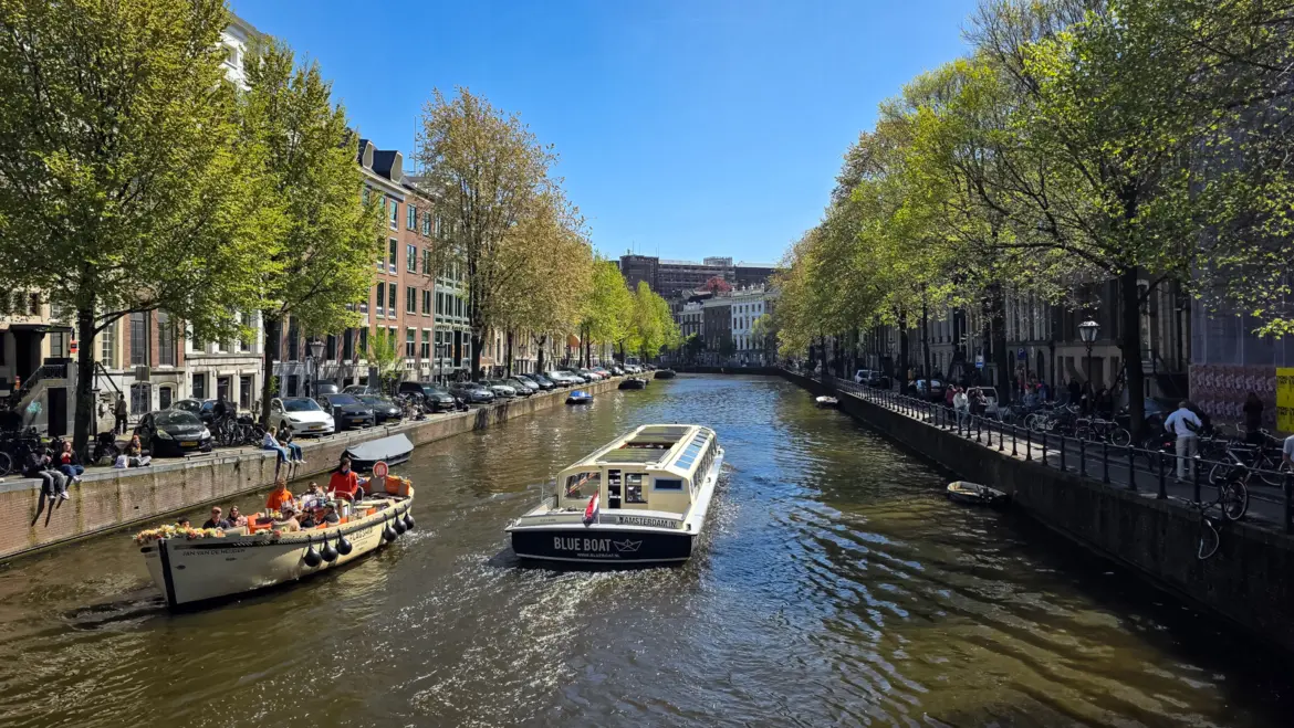 Canal boats on a tree-lined Amsterdam canal in spring sun, an open flower-decked boat with passengers in orange and a Blue Boat tour boat