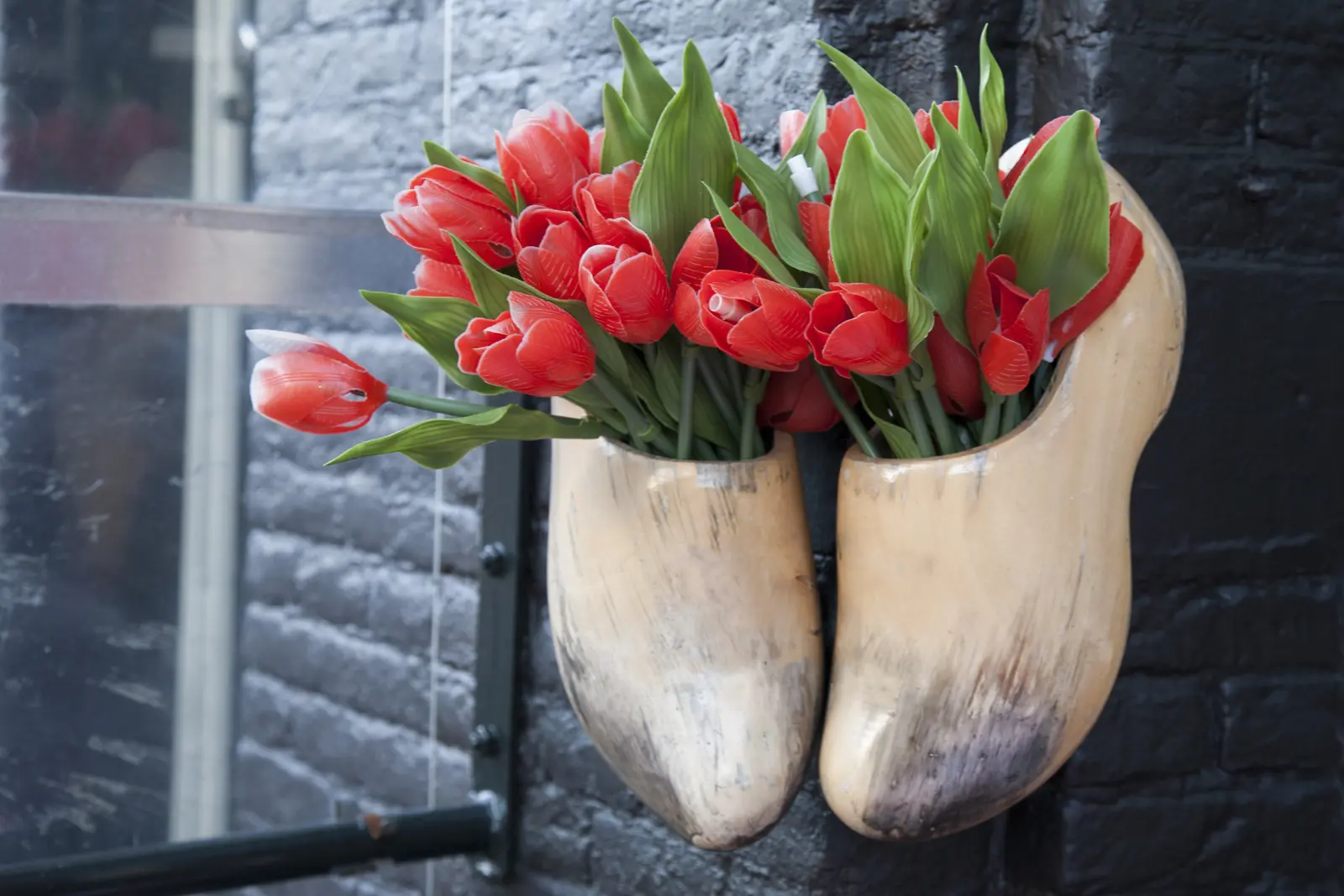 A pair of wooden clogs hung on a dark brick wall in Amsterdam, stuffed with plastic red tulips and green leaves