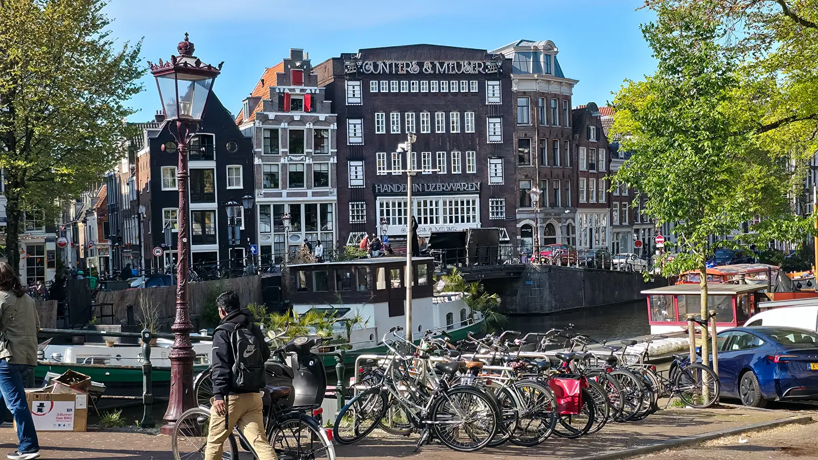Bikes racked on a canal quay opposite the Gunters & Meuser building in Amsterdam, with houseboats moored below