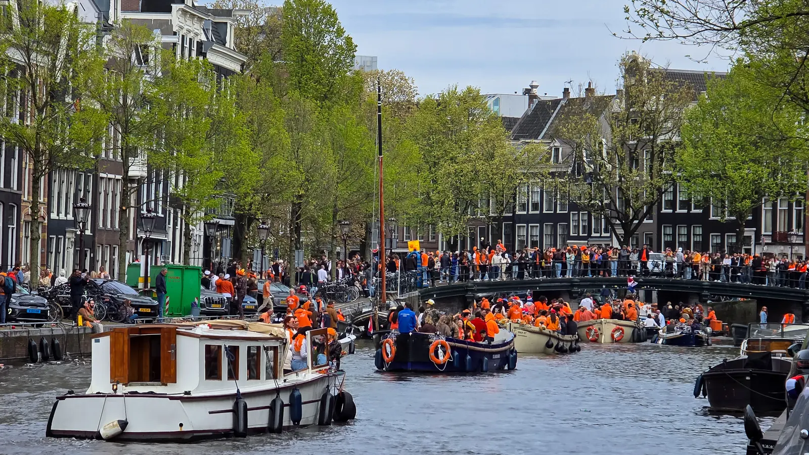 King's Day boats crowded with people in orange on an Amsterdam canal, with more crowds lining a bridge