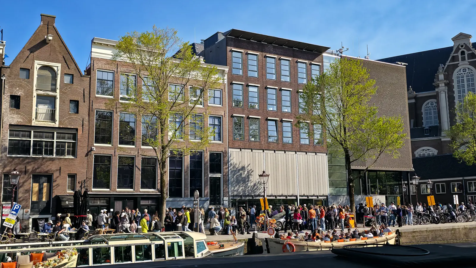 Crowds on the pavement outside the Anne Frank House on the Prinsengracht, with canal boats moored in front and the Westerkerk at right, Amsterdam