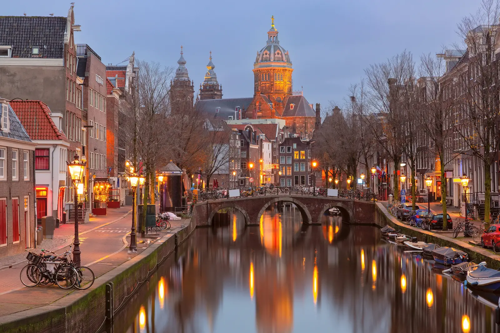 A canal and bridge near the Red Light District at dusk, with the domed Basilica of St Nicholas lit up behind