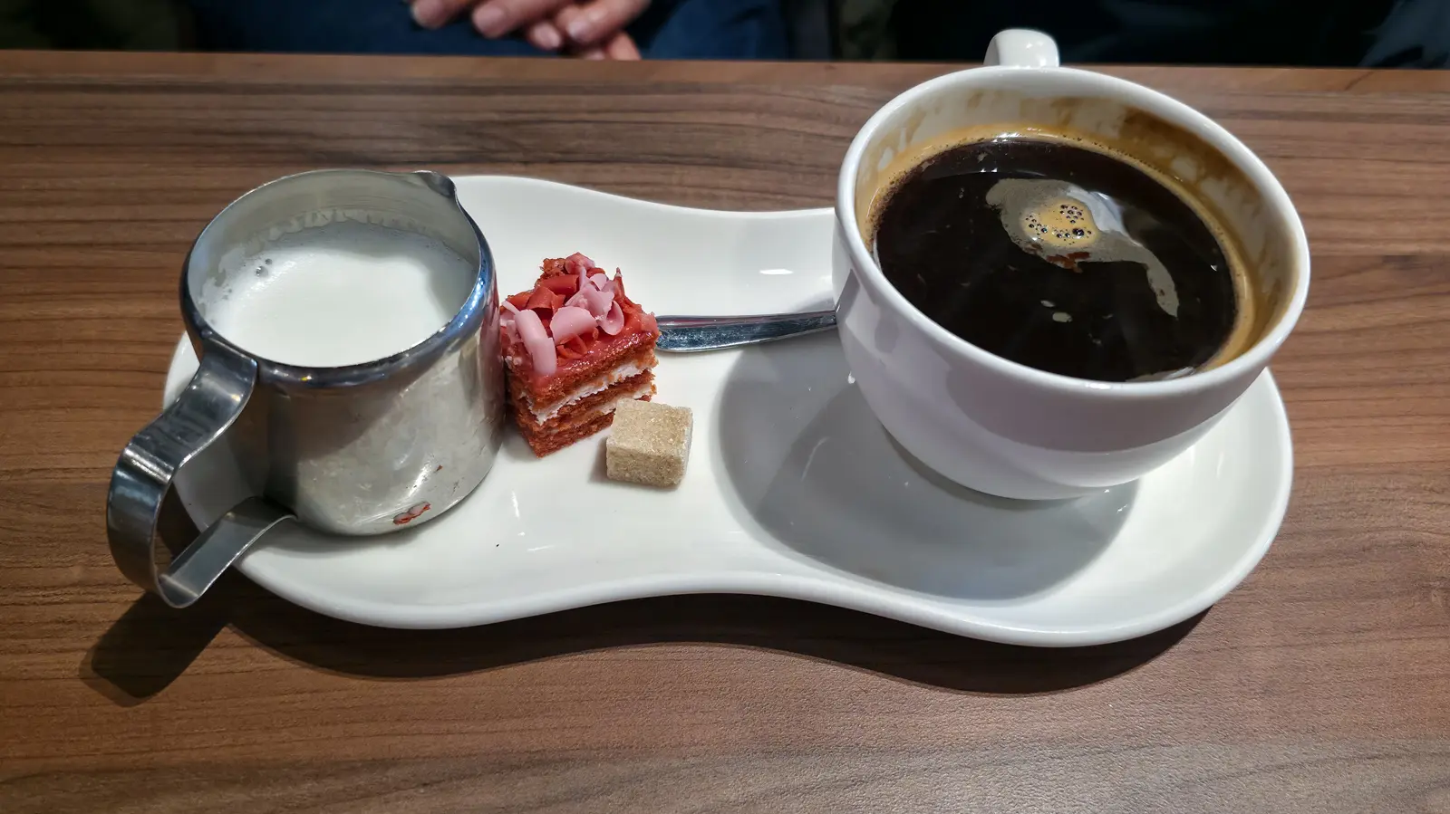 An americano with a steel jug of milk, a small layered cake bite and a brown sugar cube on a curved saucer at Café Flor, Schiphol