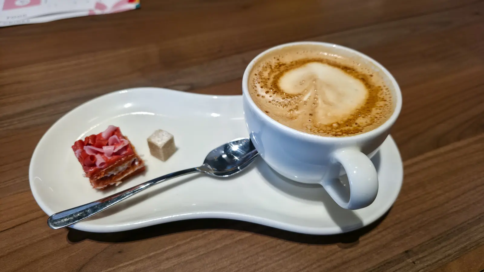 A cappuccino with a latte-art heart, a small layered cake bite and a sugar cube on a curved saucer at Café Flor, Schiphol