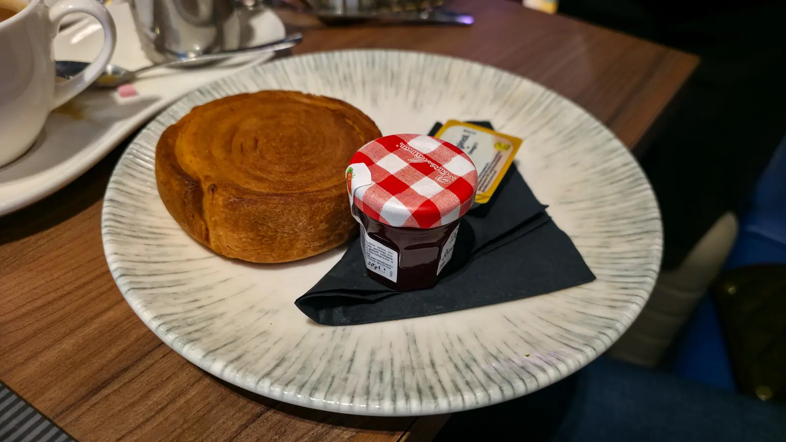 A round croissant roll with a small jar of Bonne Maman jam and a pat of butter at Café Flor, Schiphol