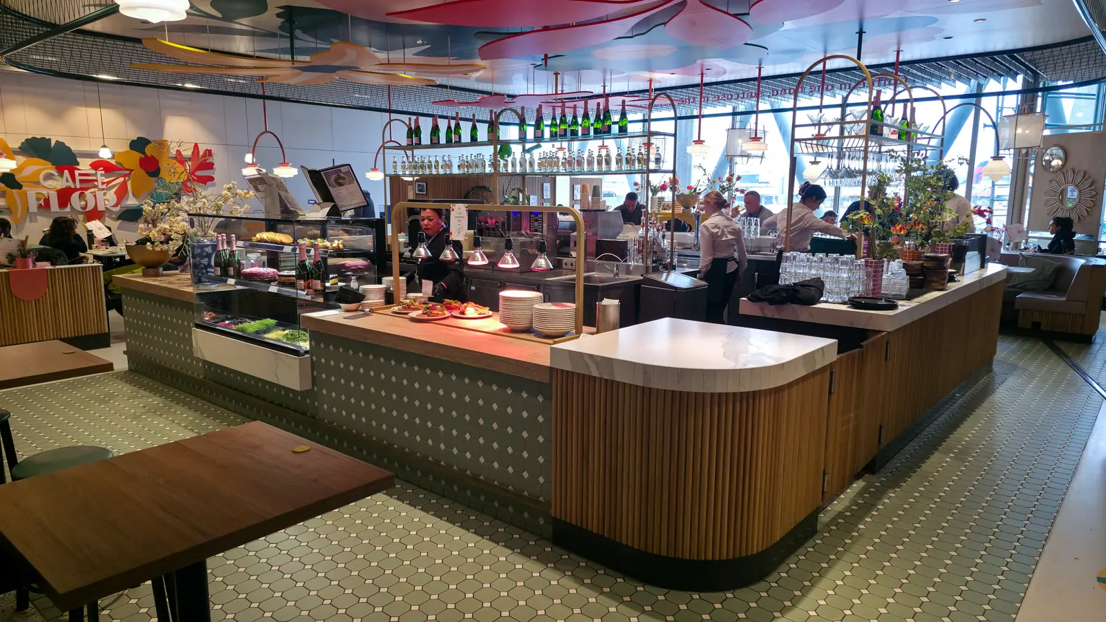 Café Flor's counter at Schiphol, with a cake display, warming lamps, champagne on the shelves and flower cut-outs overhead
