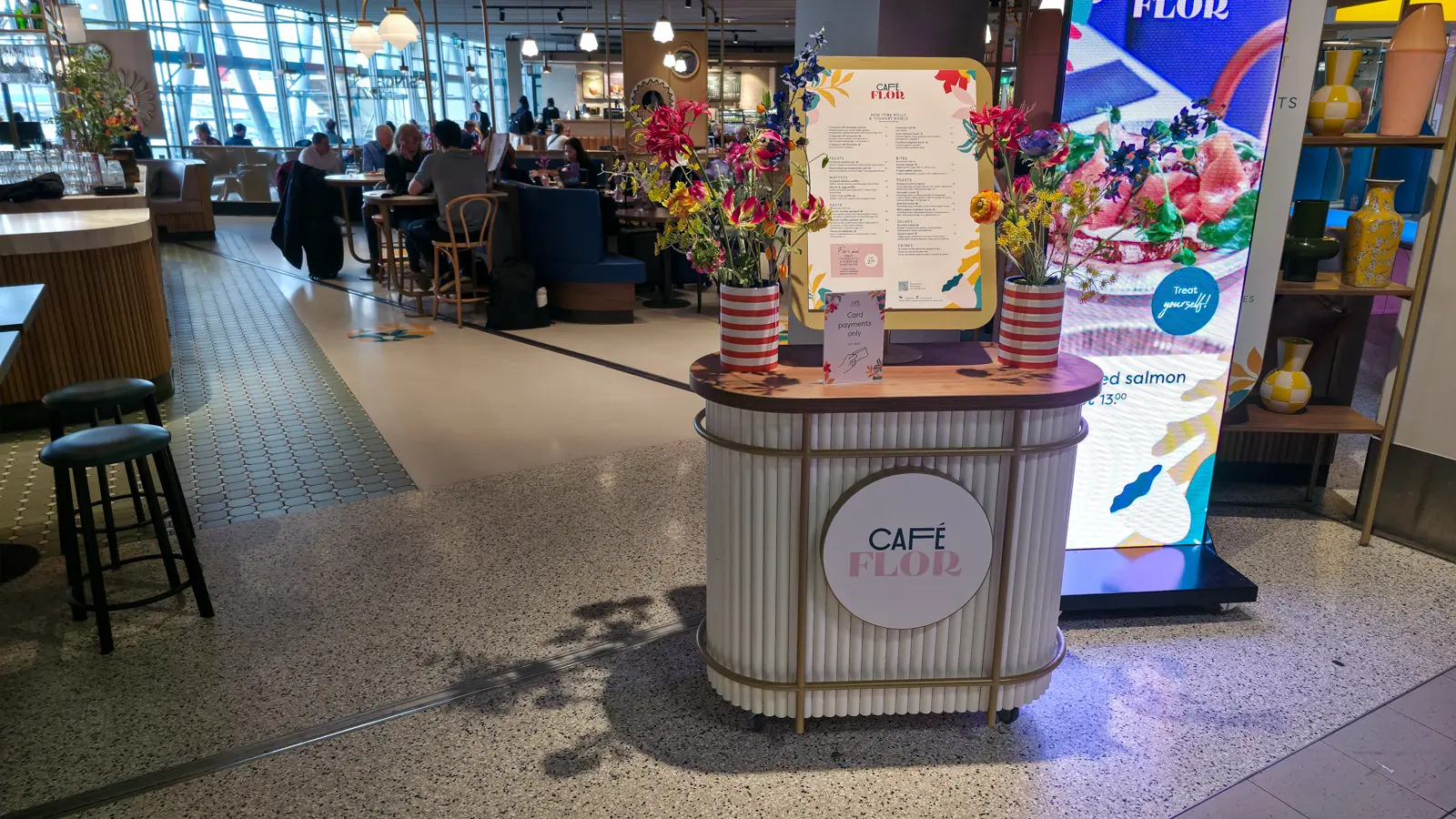 The Café Flor menu stand at Schiphol, with flowers in red-and-white striped pots and a card-payments-only sign