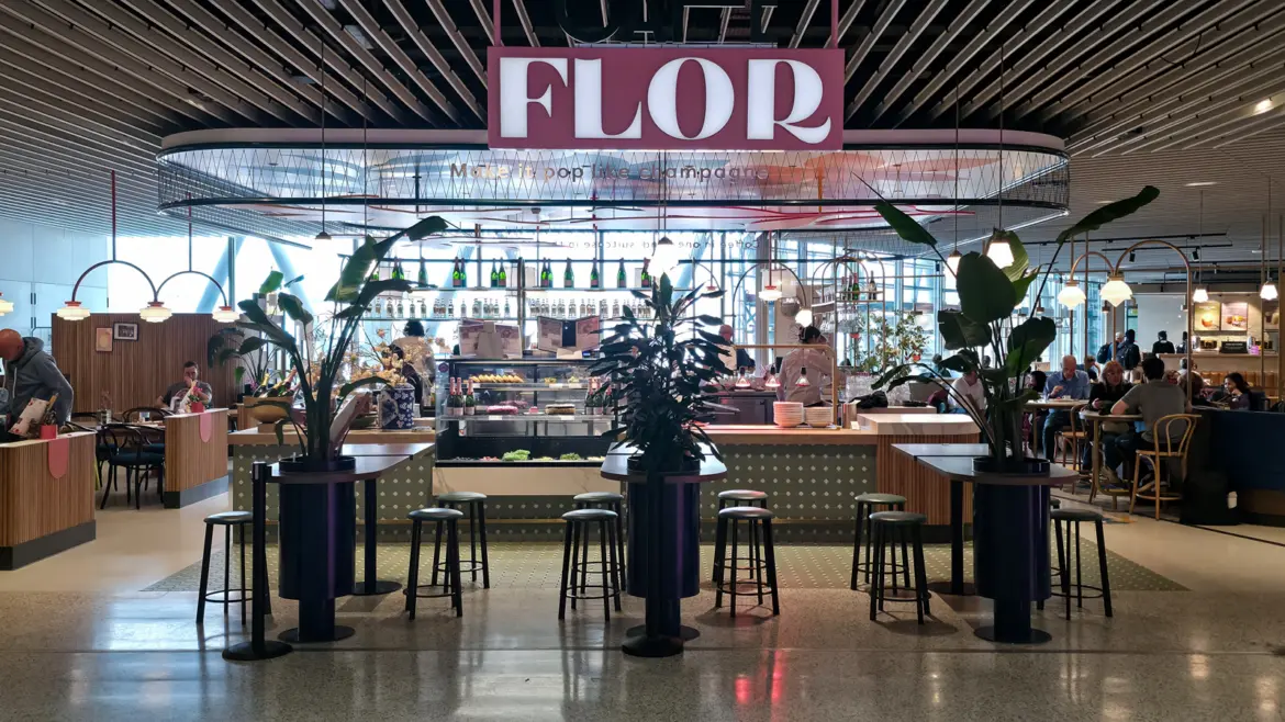 The front of Café Flor at Schiphol, with a pink FLOR sign, tall plants, bar stools and champagne bottles behind the counter
