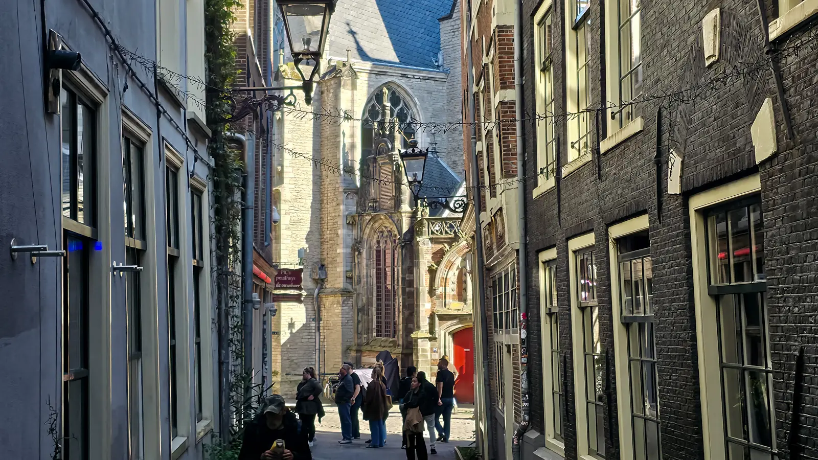 People gathered at the end of a narrow De Wallen alley, with lanterns overhead and the Oude Kerk's Gothic facade behind
