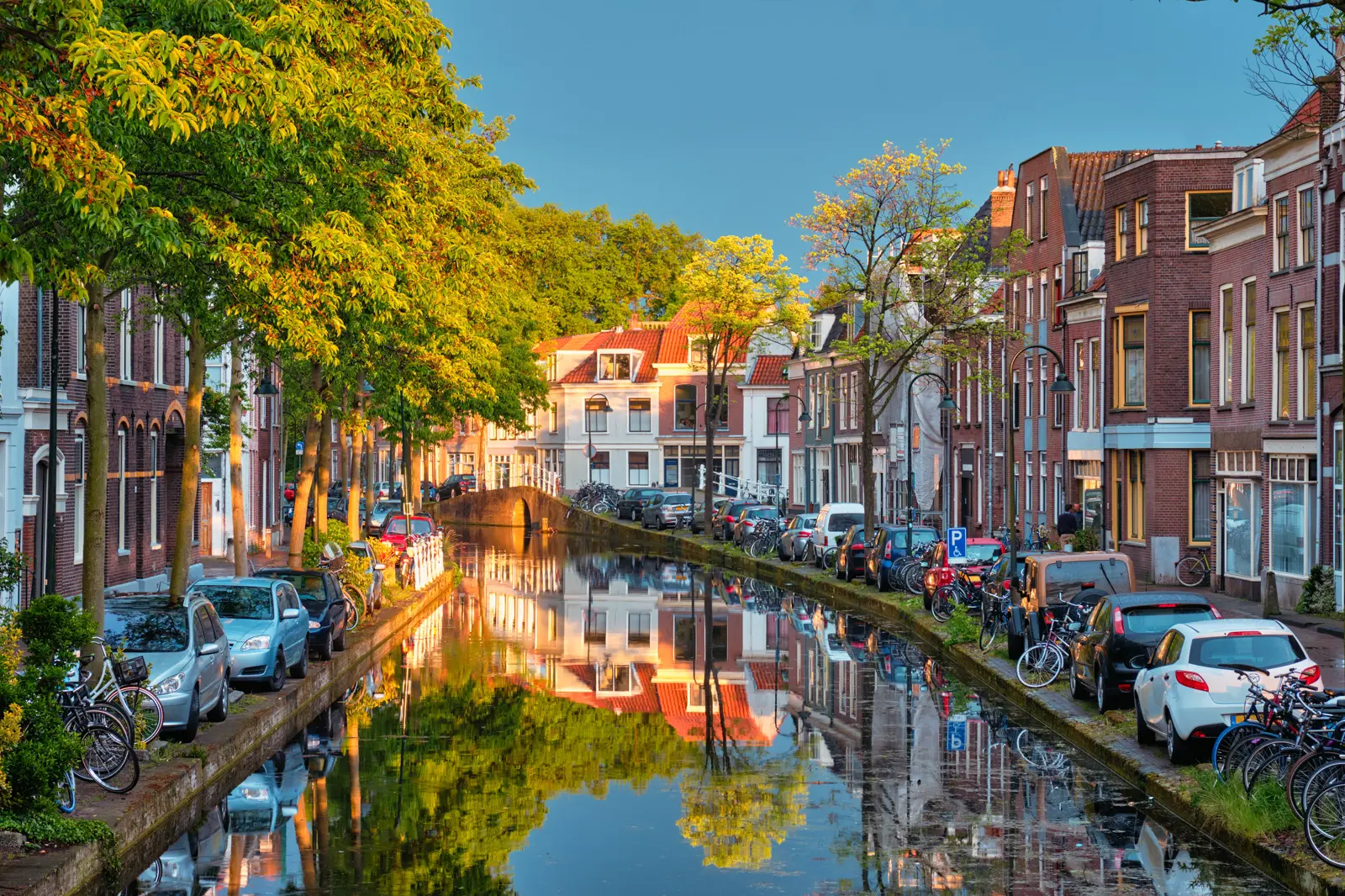 A tree-lined canal in Delft at sunset, with brick houses, parked bikes and a humped bridge reflected in the water