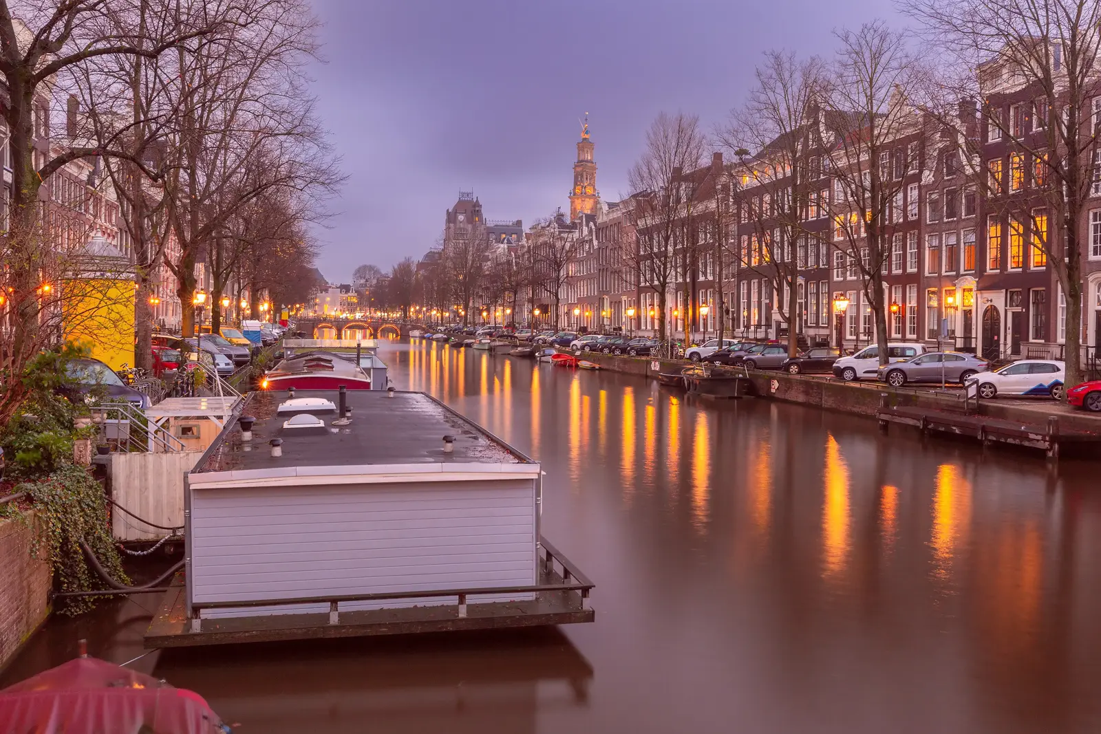 Houseboats moored along a tree-lined Amsterdam canal at dusk, with a church tower lit up at the far end
