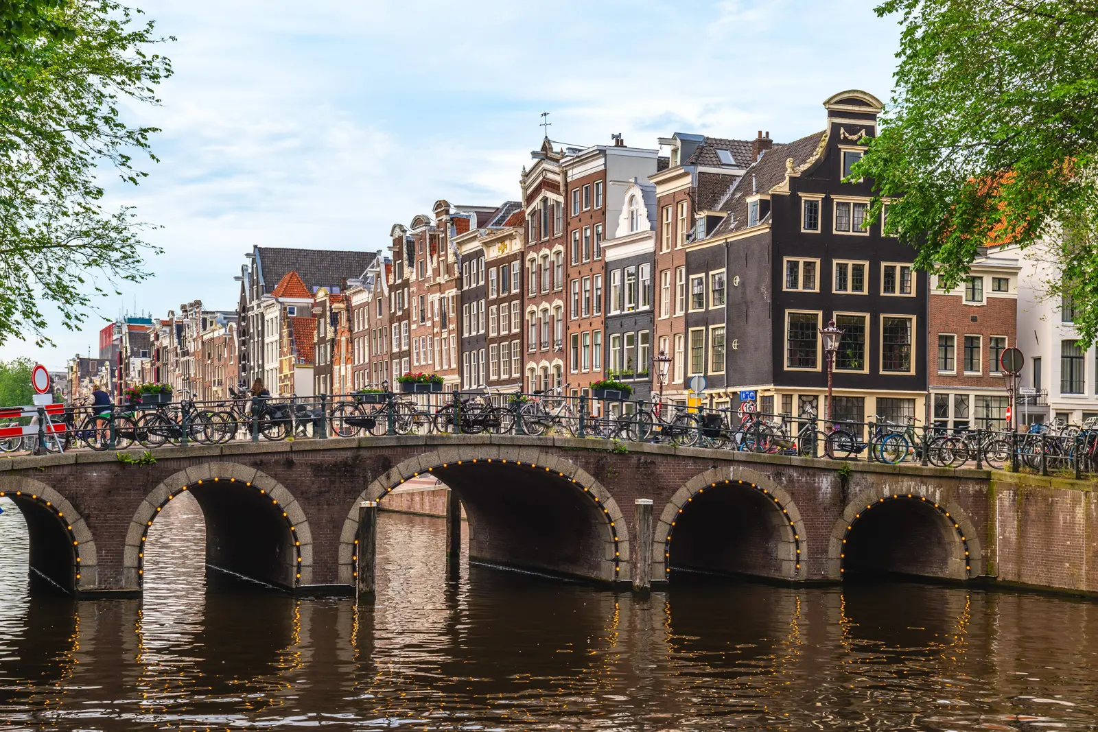 An arched brick bridge lined with parked bikes, with tall gabled canal houses behind, in Amsterdam's Canal District