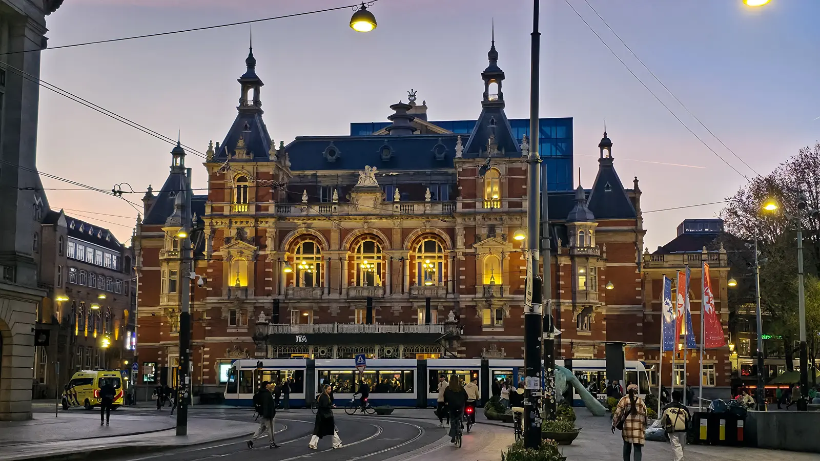 A tram passing the illuminated International Theater Amsterdam (Stadsschouwburg) on the Leidseplein at dusk, with people crossing in front, Amsterdam
