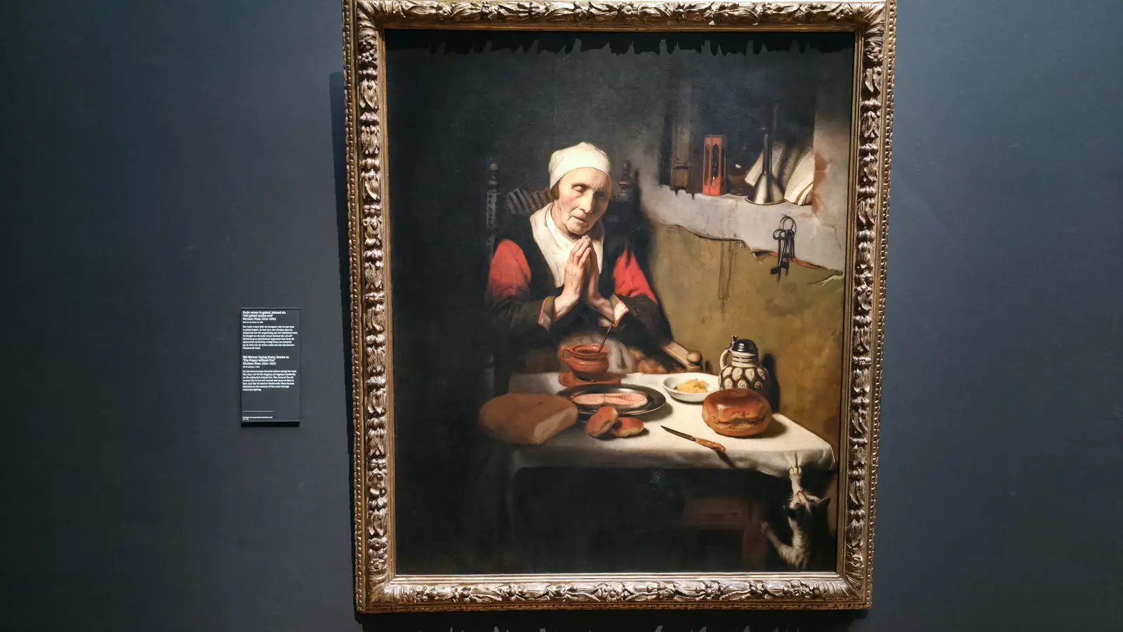 Nicolaes Maes' Old Woman Saying Grace, an elderly woman praying over bread and fish with a cat clawing the tablecloth at lower right, Rijksmuseum Amsterdam