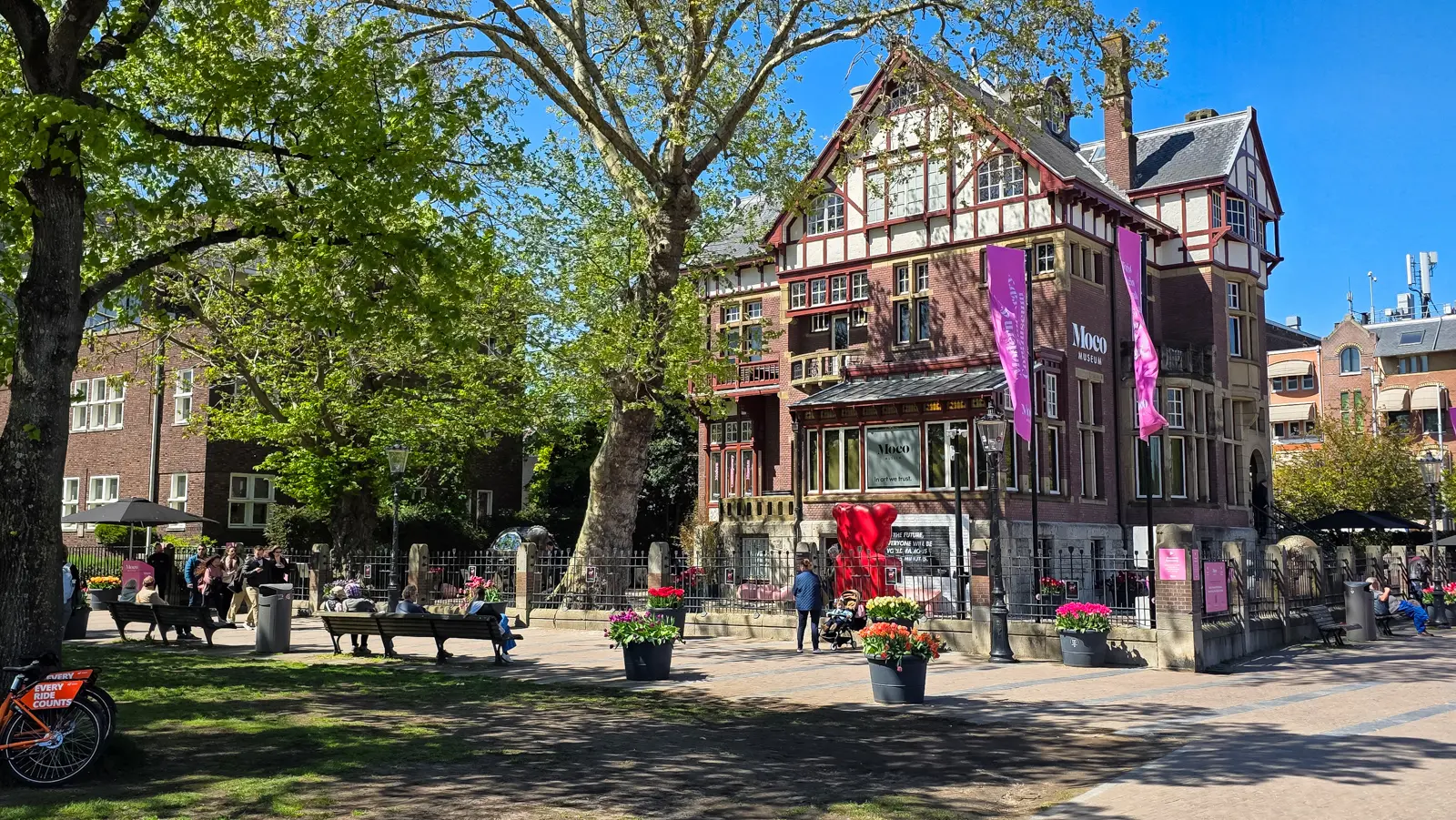 Moco seen from the park side under plane trees, with benches, tulip planters and a large red sculpture by the fence