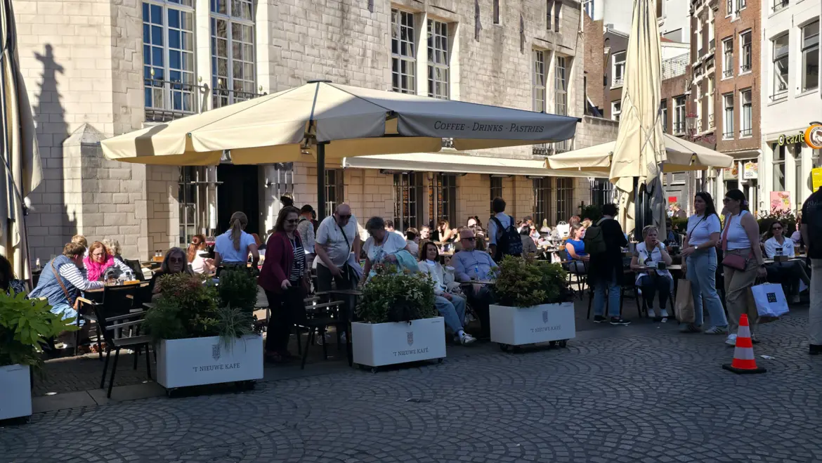 The packed terrace of 't Nieuwe Kafe on Dam Square, Amsterdam, with white planters and cream umbrellas against the pale stone of the Nieuwe Kerk