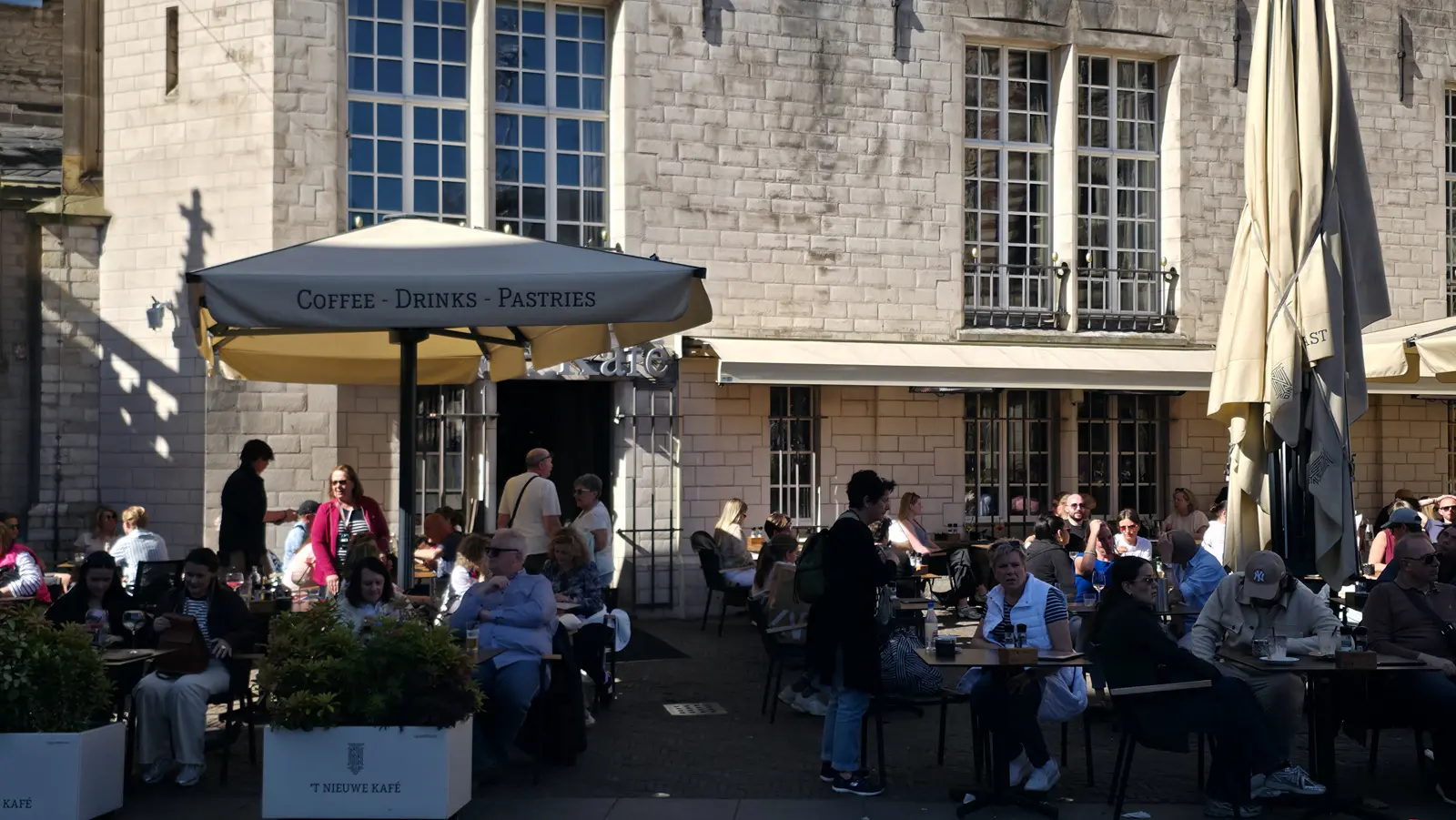 Diners under a Coffee, Drinks, Pastries umbrella outside the pale stone facade of 't Nieuwe Kafe, Amsterdam