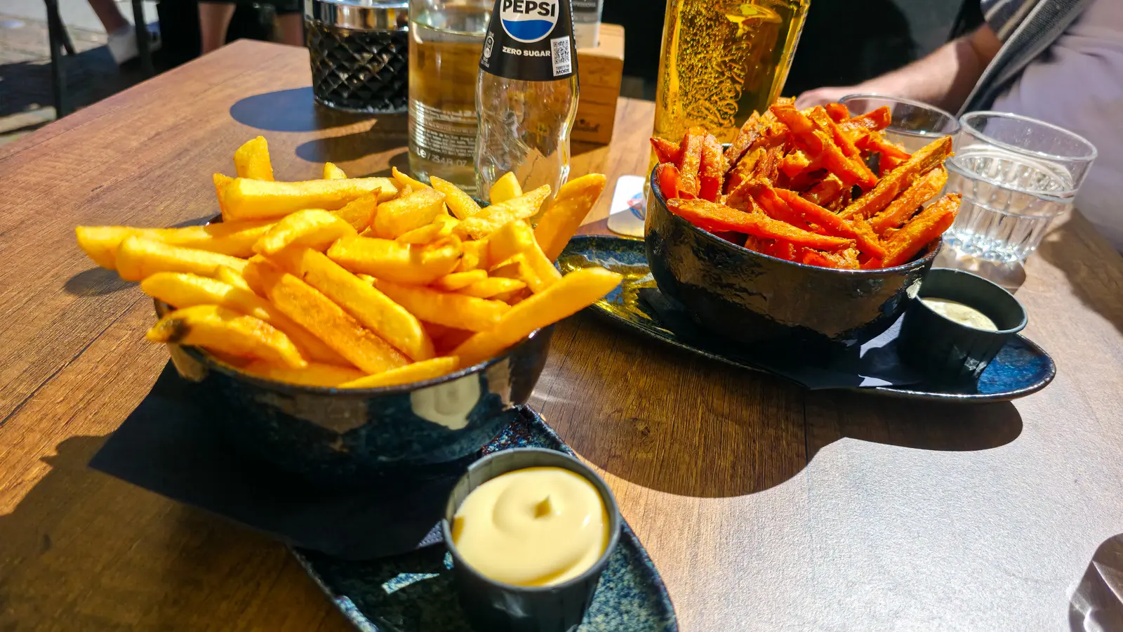A bowl of regular fries in the foreground and sweet potato fries behind, on a table at 't Nieuwe Kafe