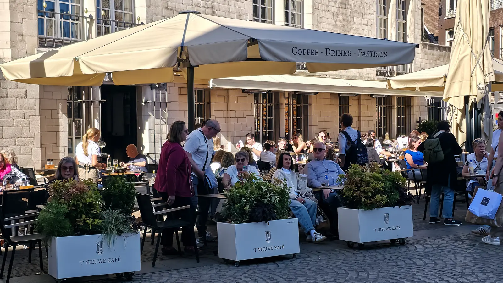 A packed terrace at 't Nieuwe Kafe on Dam Square, with greenery in white planters and cream Coffee-Drinks-Pastries umbrellas